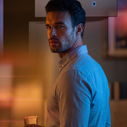 A man in a light blue button-down shirt turns to look over his shoulder with a serious expression, illuminated by dramatic warm orange and cool blue lighting inside a modern home.